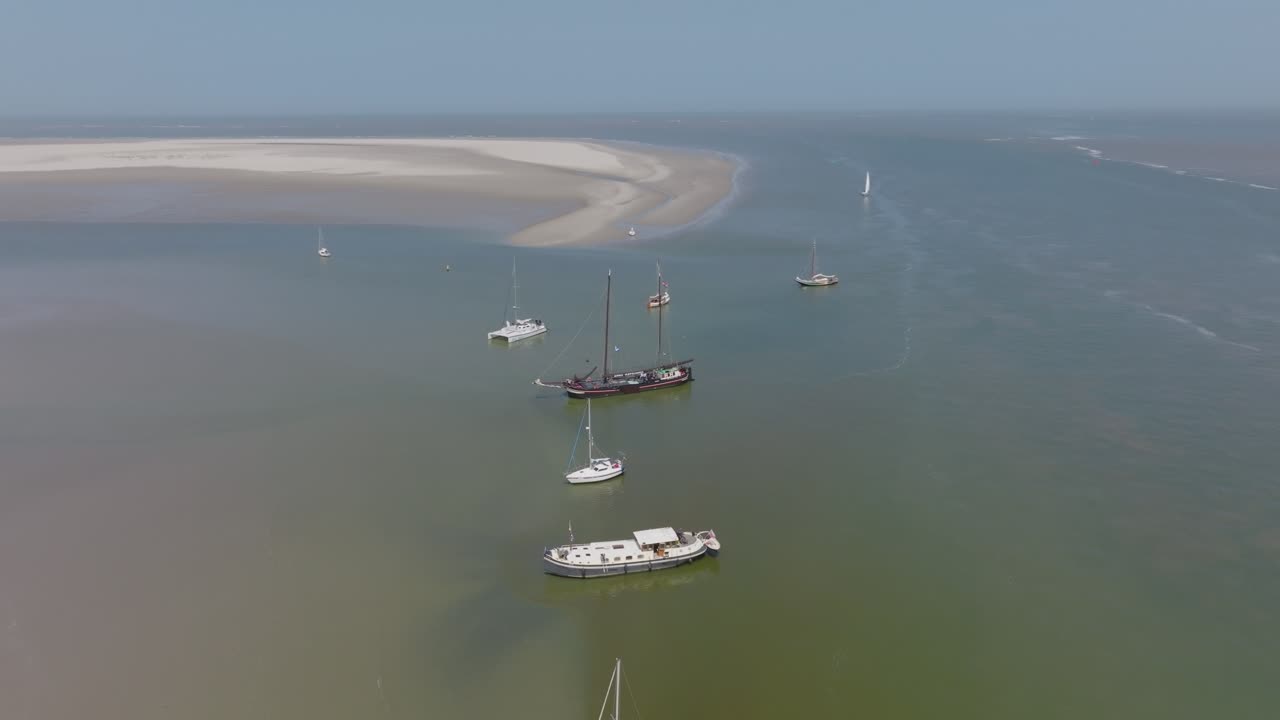 Wide aerial of several boats anchored over green water beside pale sandbars at low tide; oblique high view, daylight. Wadden Sea