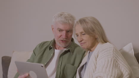 senior couple sitting on sofa at home using digital tablet 1