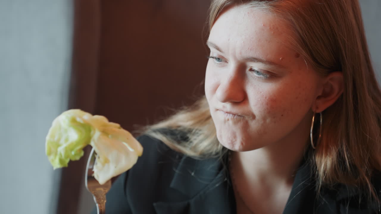 Femme blanche inspectant une feuille de laitue sur une fourchette. Gros plan d'une critique culinaire milléniale dans un café avec des boucles d'oreilles créoles et une peau tachetée de rousseur, lumière douce de la fenêtre, expression passant de la curiosité au dégoût, léger mouvement de tête