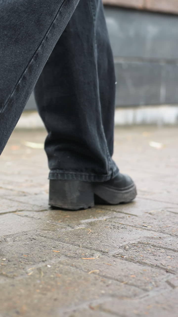 Close up of human leg wearing black trousers and black boots walking slowly on outdoor paved path during calm overcast day, capturing foot movement and subtle step motion
