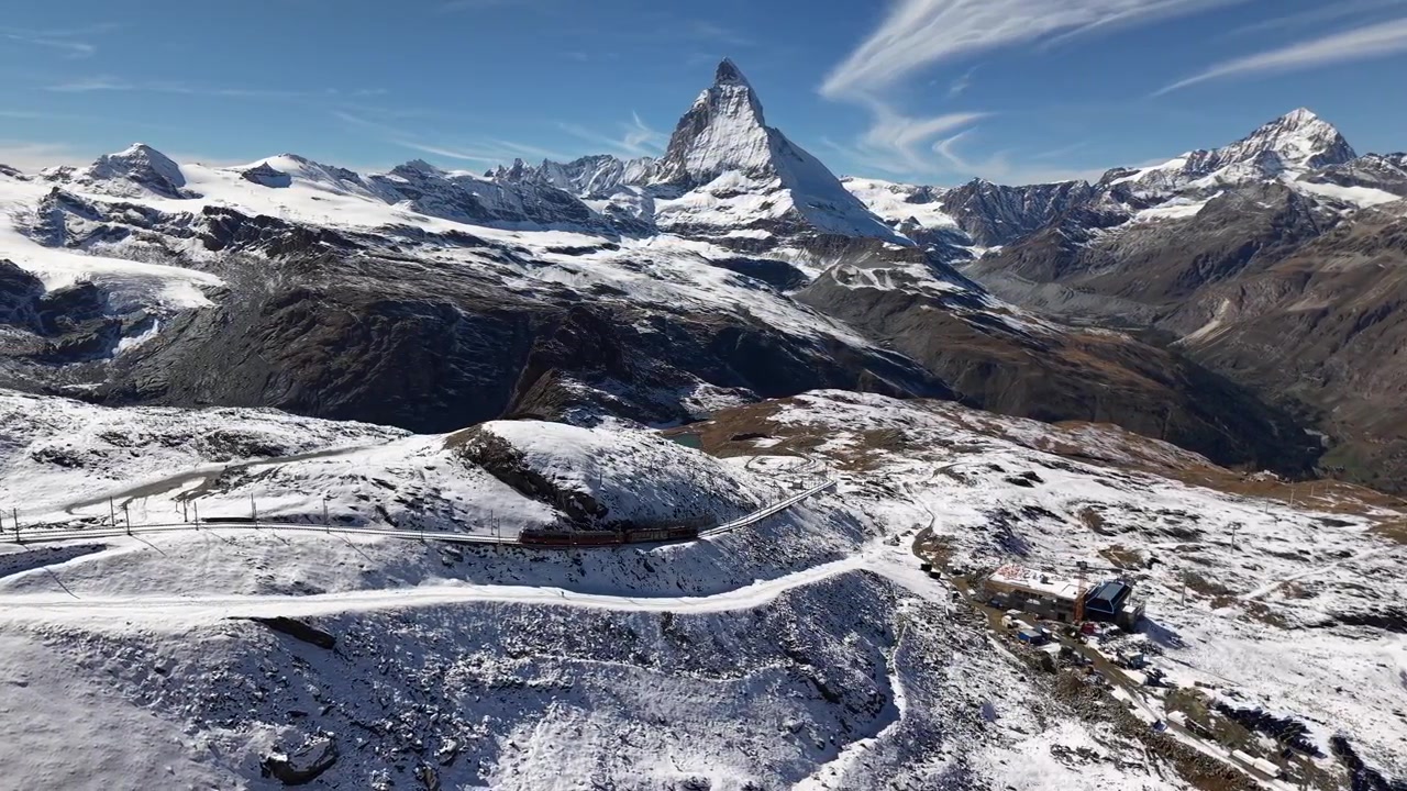 スイスアルプスのツェルマット、ゴルナーグラート鉄道沿いを空撮。雪の降る秋の日にマッターホルンを背景に列車が下っていく様子