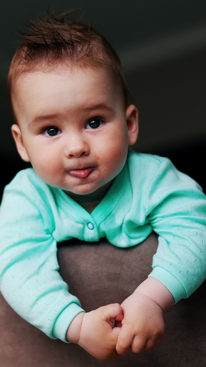 Lovely toddler looking around standing peacefully on the chair. Adorable baby showing tongue and smiling. Low angle view. Vertical video