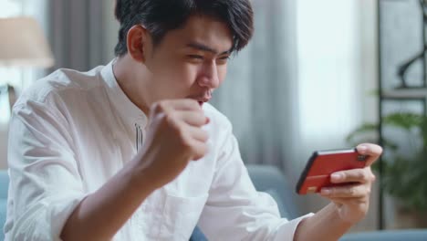 close up side view of asian man celebrating winning game on smartphone while sitting on sofa in the living room