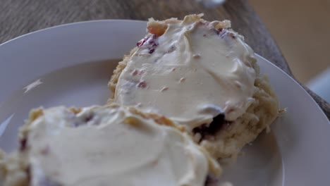 Person-spreading-whipped-thick-cream-over-homemade-traditional-baked-jam-scone-with-silver-metal-nice-on-white-plate-and-timber-table-in-rustic-setting-for-afternoon-tea