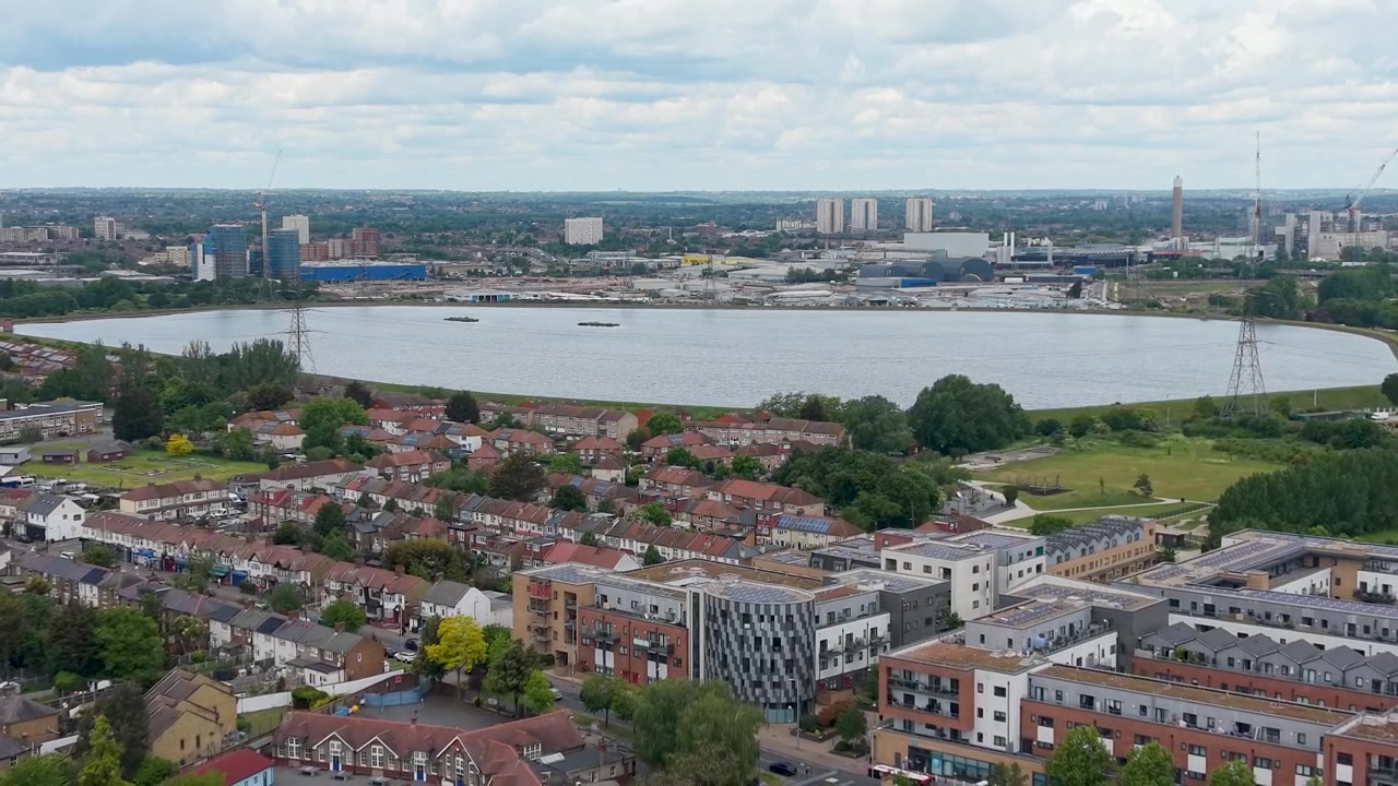 Ripresa aerea con drone in allontanamento dal bacino idrico di Banbury verso Walthamstow da South Chingford, che mostra tetti, aree verdi, le acque della Lea Valley, tralicci e lo skyline di Londra in lontananza sotto le nuvole.