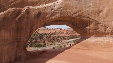 Drone-Flying-On-The-Hole-Of-Wilson-Arch-Revealing-The-US-Highway-191-In-The-Distance-On-A-Sunny-Day-In-Moab,-Utah,-USA