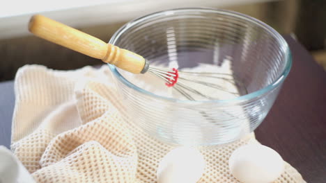 Whisk-in-glas-bowl.-Closeup.-Eggs-on-kitchen-table.-Hand-throws-whisk-in-a-bowl