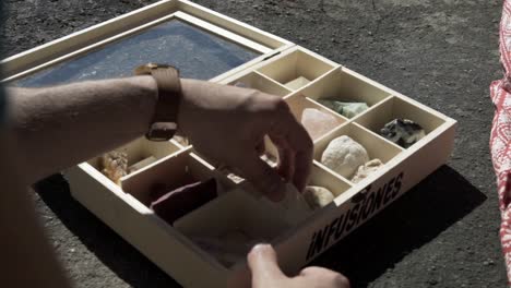 una caja de forma cuadrada contiene numerosos tipos de rocas con atributos específicos, y el hombre está mirando cada una