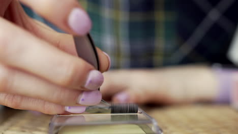 mujer con uñas rosas sacando pestañas de una caja de plástico con pinzas para practicar el procedimiento cosmético, disminuyendo el número de pestañas del cliente