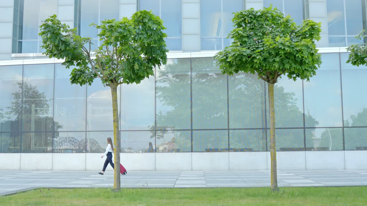 Girl walking with red suitcase in front of glass building, passing two maple trees