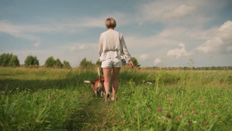 dog trainer walks through open grassy field with her dog playfully dragging her forward, background features vast landscape of greenery and trees under clear blue sky