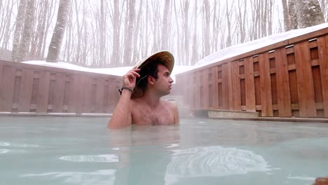 foreign male tourist wearing straw hat inside open air onsen with snow in japan