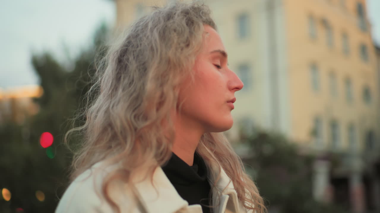 Side view of calm lady walking looking unhappy holding white umbrella glancing around with blurred background of trees city lights and residential buildings under soft evening sky