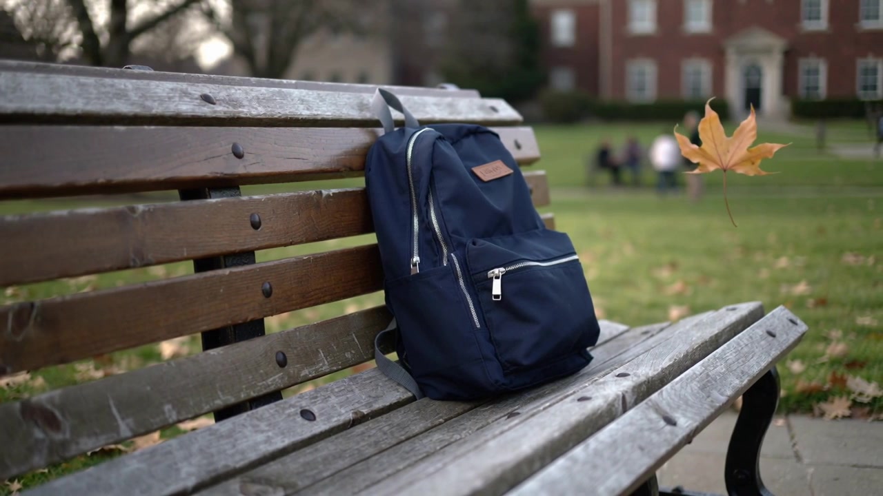 Breeze sending brown maple leaf drifting across park bench, passing navy backpack, copy space