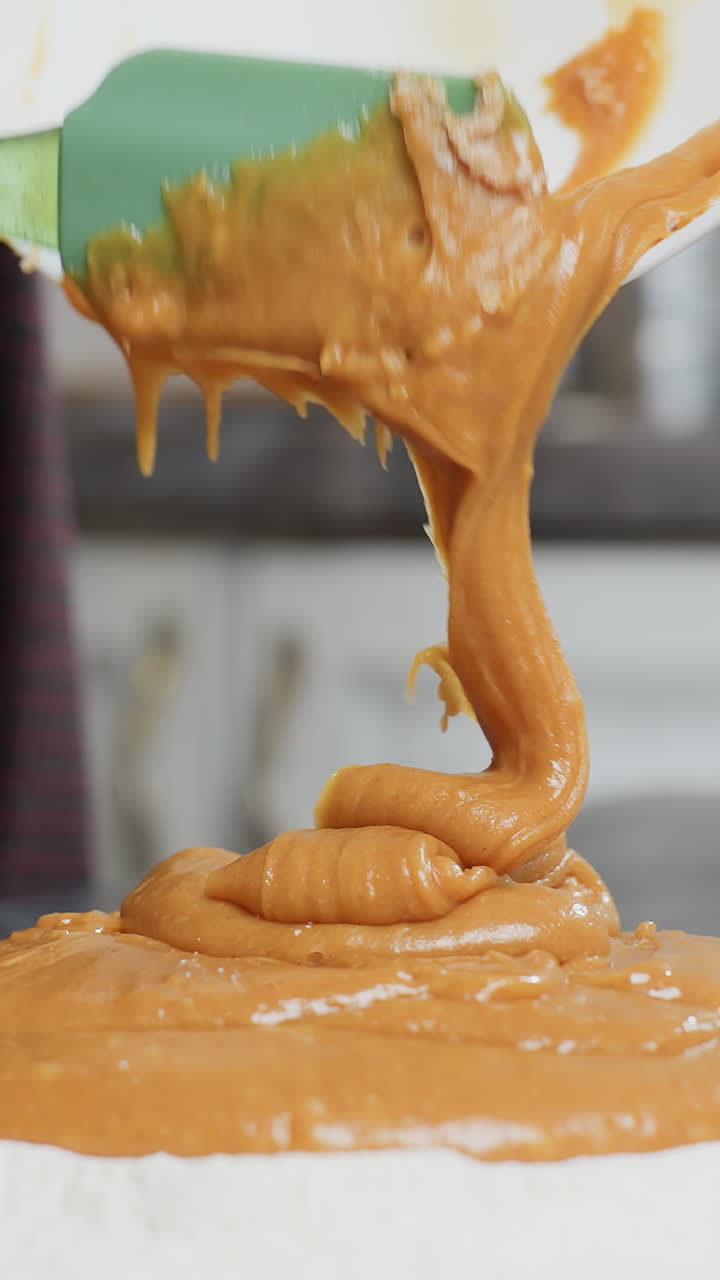 녹색 스파라를 사용하여 카운터에 두꺼운 카라멜 색의 반죽을 밀가루로 부는 여성의 클로즈업, 부드럽고 풍부한 혼합물이  그에서 흐릅니다.