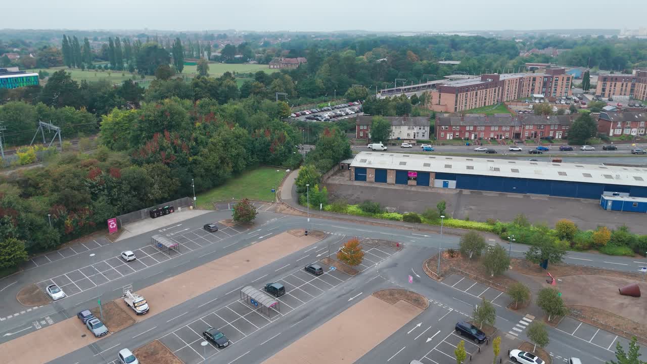 Aerial view of an urban area with parking lots, buildings, and roads amidst greenery under an overcast sky. The scene includes residential and commercial structures