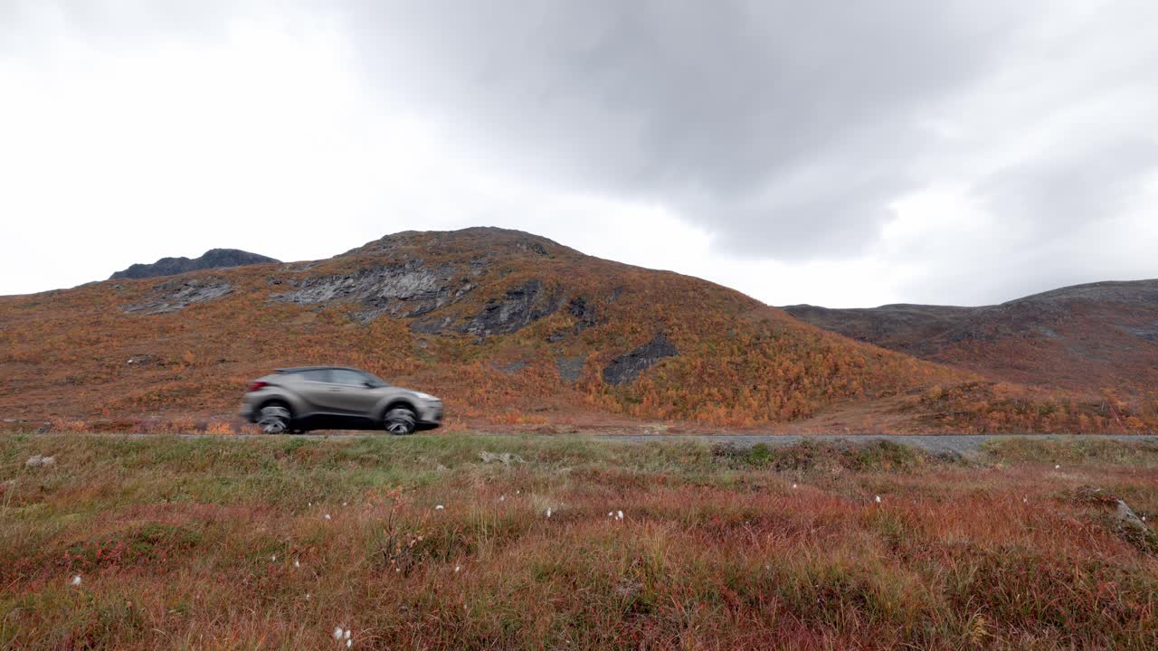Un SUV argentato percorre una remota strada di montagna attraverso uno splendido paesaggio autunnale in Norvegia. Colline spettacolari coperte da un fogliame autunnale dorato-arancio sotto un cielo coperto creano una scena suggestiva e atmosferica