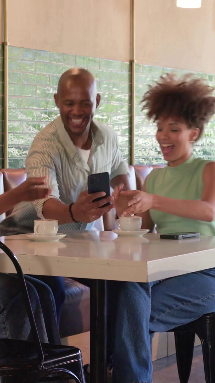 Vertical video: Arriving man with cup hugging woman in green top leaning showing phone at cafe