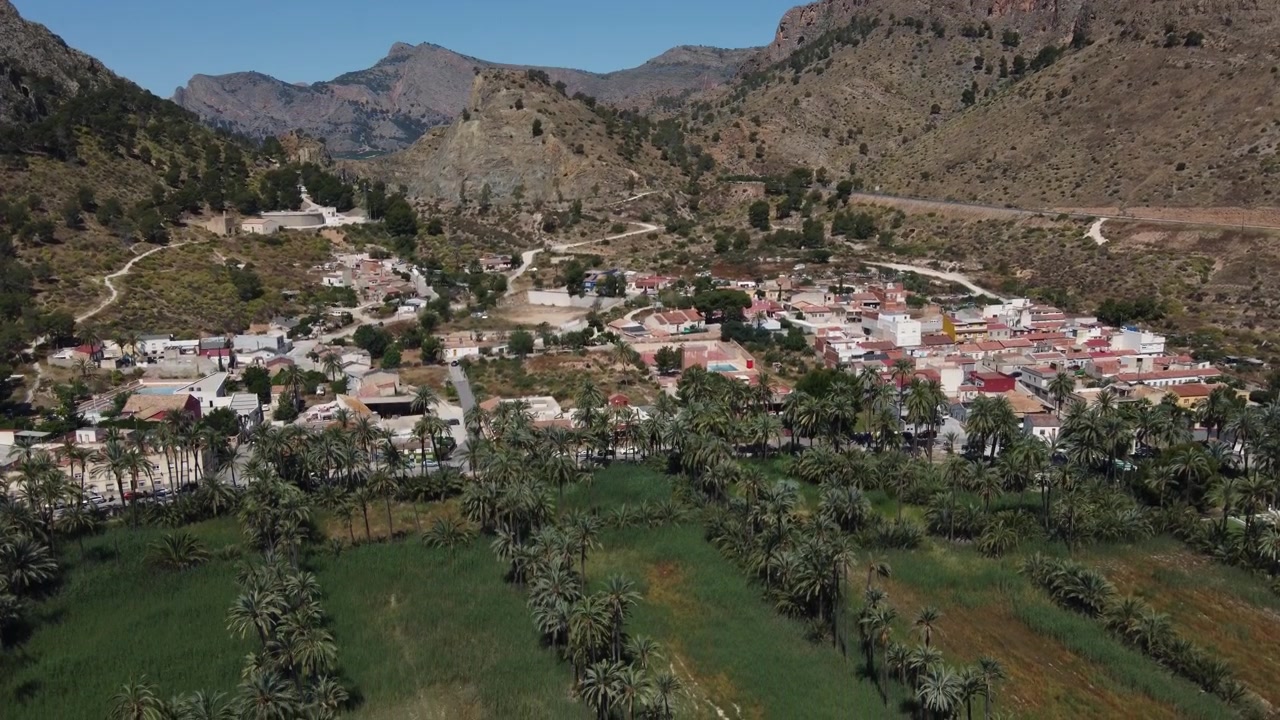 Ripresa aerea in discesa con drone del villaggio di San Antón, immerso nel lussureggiante palmeto di Orihuela, circondato da aride montagne desertiche ad Alicante, Spagna