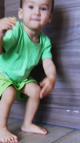 curious toddler boy sits on the floor holding some food in his hand. cute baby stands up and approaches camera. low angle view. vertical video