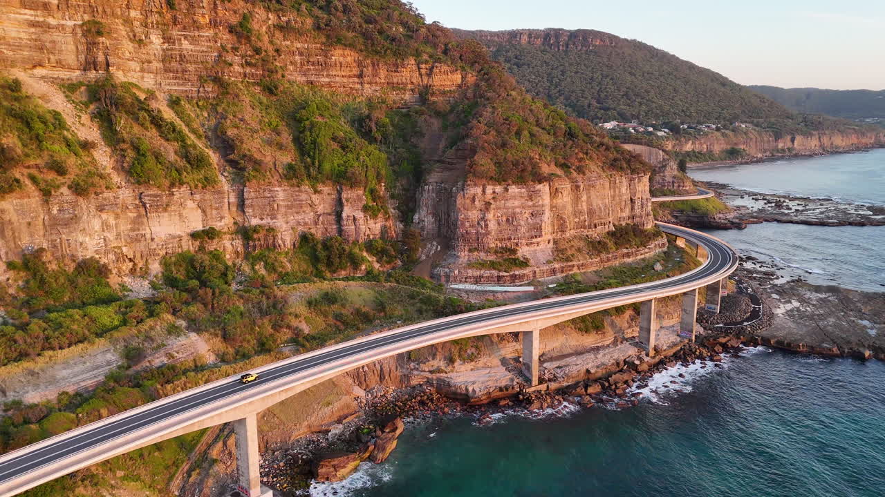 Epic aerial view of a car on the Sea Cliff Bridge, revealing the winding highway perched above the rugged Australian coastline. Wide angle drone footage, golden hour.
