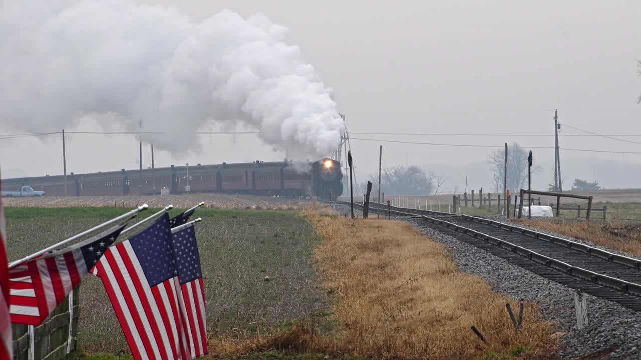 una vista a largo plazo de una línea de banderas americanas ondeando suavemente en una valla junto a las tierras de cultivo mientras un tren de pasajeros de vapor dobla una curva, soplando humo en un día de invierno