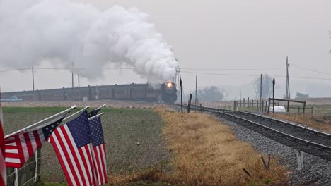 A-Long-View-of-a-Line-of-Gently-Waving-American-Flags-on-a-Fence-by-Farmlands-as-a-Steam-Passenger-Train-Rounding-a-Bend,-Blowing-Smoke-Approaches-on-a-Winter-Day