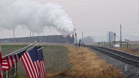 una vista a largo plazo de una línea de banderas americanas ondeando suavemente en una valla junto a las tierras de cultivo mientras un tren de pasajeros de vapor dobla una curva, soplando humo en un día de invierno