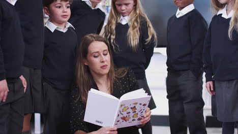 primary school kids standing behind their teacher, sitting on the floor in class reading them a book