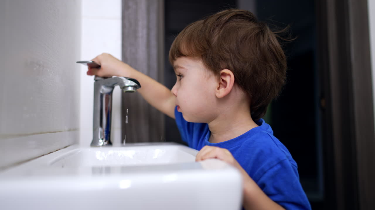Adorable kid watching the water flow. Lovely baby puts his hand under the water and moves the tap. Close up.