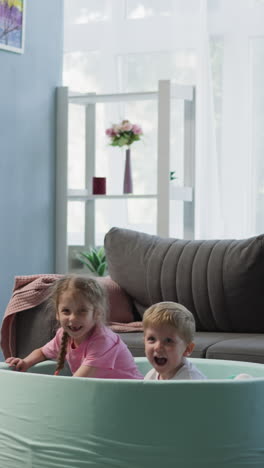 little brother and sister jump into light blue dry pool with colorful plastic balls in living room. joyful children laugh having fun together at home