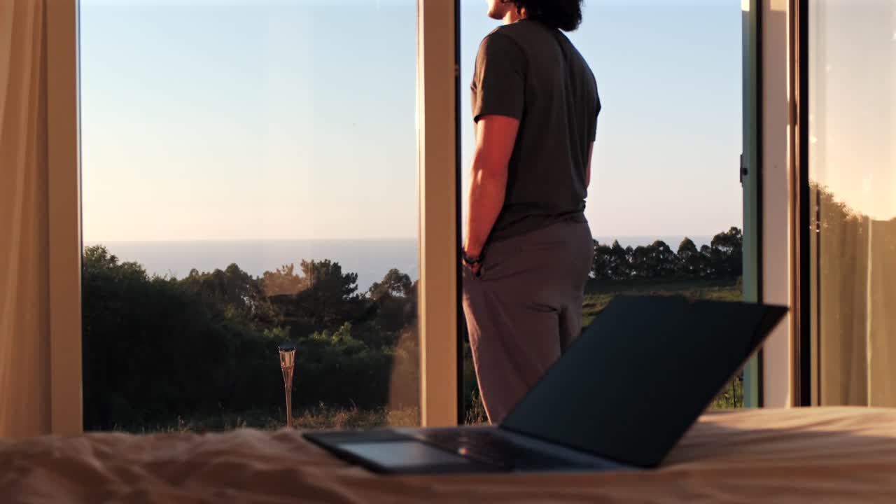 Man standing by a window, laptop on bed by the ocean