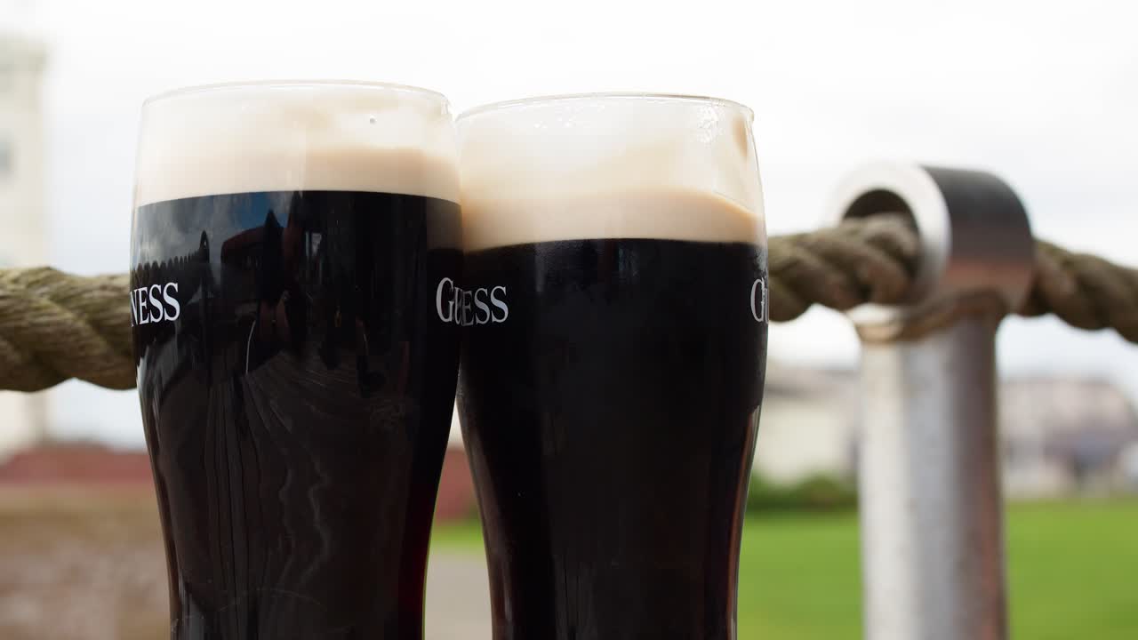 Two people clink pints of dark beer outdoors, natural daylight, shallow depth of field