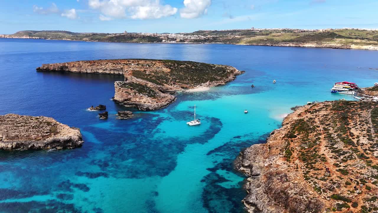 Droneoptagelse over Den Blå Lagune i Comino, Malta, der viser klippekystlinje og levende turkisblåt vand, med lagdelte klipper og roligt hav under klart sollys.
