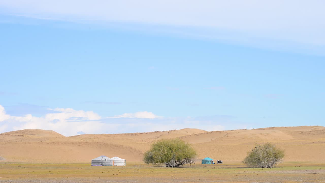 In the vast, arid landscape of the Durgun Nuur desert, a small encampment of traditional gers stands against the sand dunes. A scene of human resilience in a harsh environment