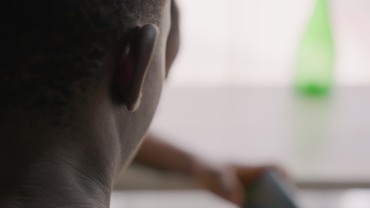 Black man checking phone screen from rear profile closeup, neck and ear in shallow focus with bokeh bottle on table, finger tapping to reply, quiet introspective mood suggesting waiting for romantic