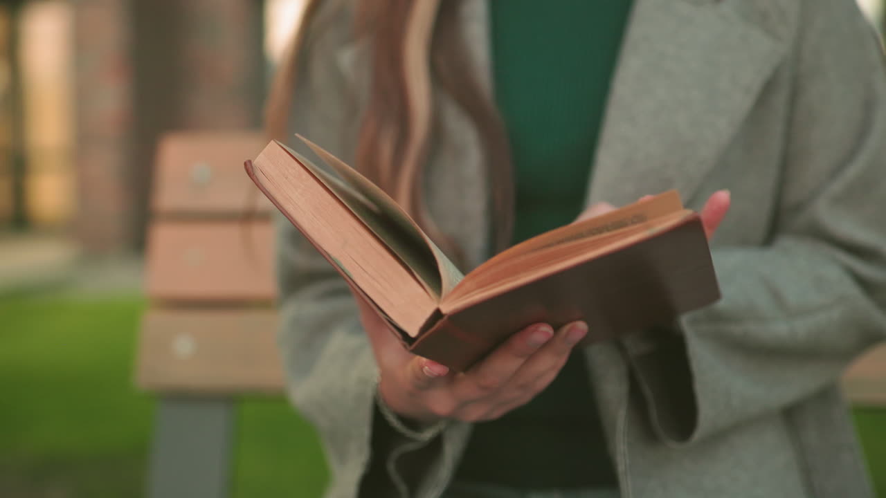 Close up of woman holding and opening book outdoors, fingers flipping pages with visible text, emphasizing learning, study, and thoughtful lifestyle