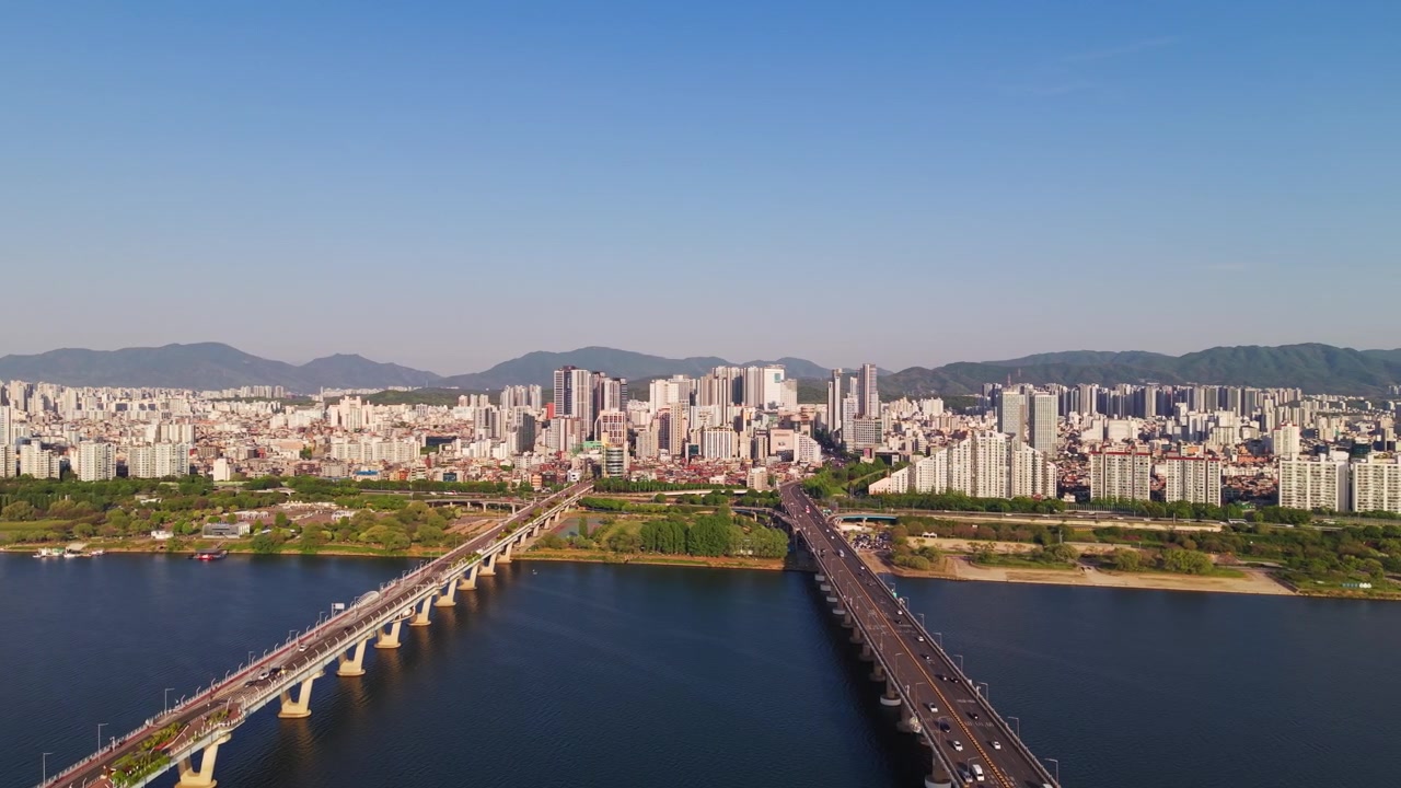 Una prospettiva aerea sui ponti Gwangjin e Cheonho che attraversano il fiume Han rivela il denso paesaggio urbano di Seul, il parco fluviale e lo sfondo montuoso.
