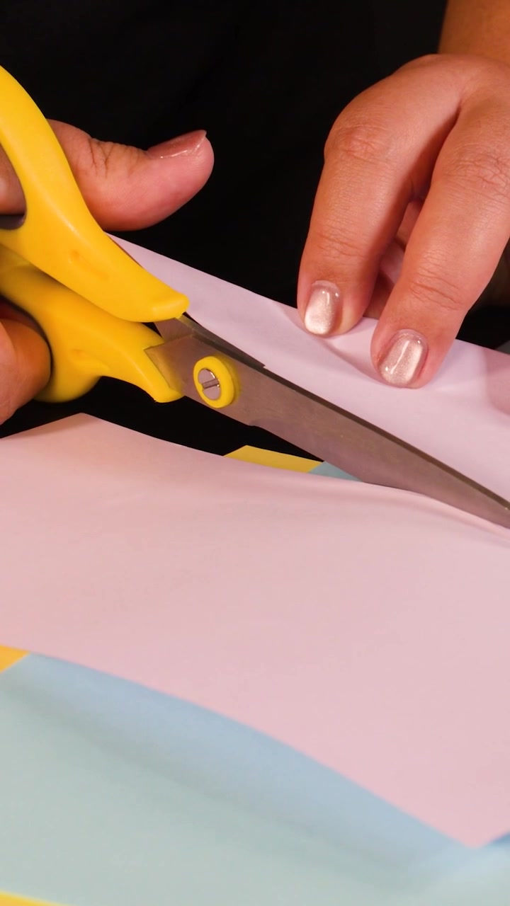 Una toma vertical de cerca que muestra manos cortando con precisión papel rosa usando tijeras de color amarillo brillante.
