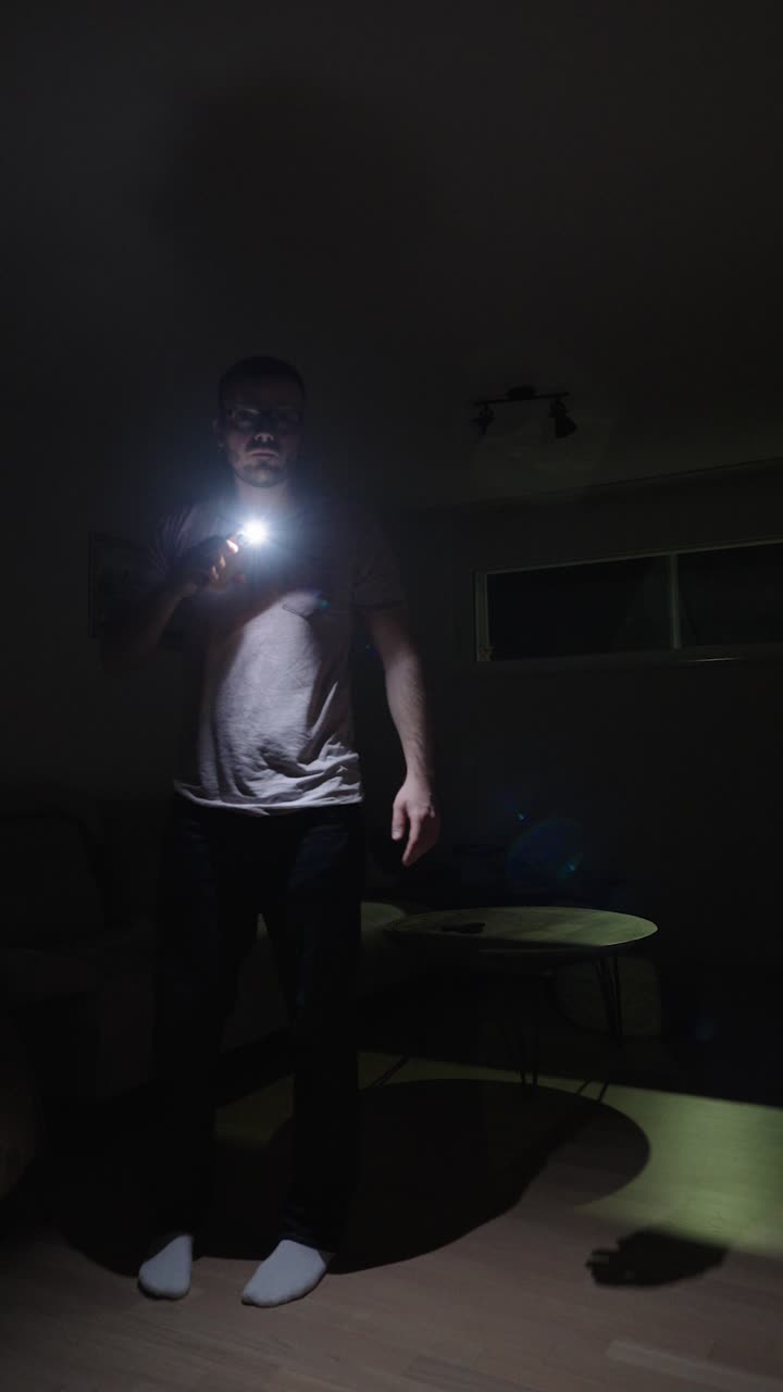 Vertical shot of a man standing in a dark living room during a power outage, using his smartphone as a light source, showing blackout, home emergency, and realistic nighttime lifestyle