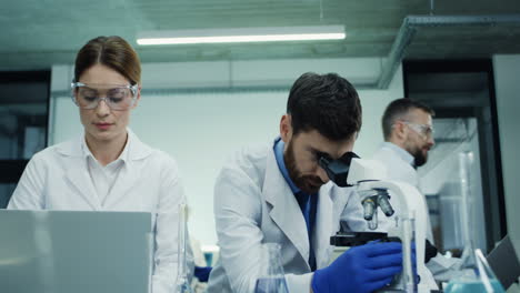 Portrait-Of-The-Two-Laboratory-Scientists,-Man-And-Woman,-Talking-While-He-Working-At-The-Microscope-And-She-At-The-Laptop-Computer