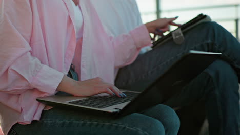 close-up of woman in pink top and jeans, polished nails typing on laptop, showing something on her tablet to her focused colleague next to her who is also typing swiftly