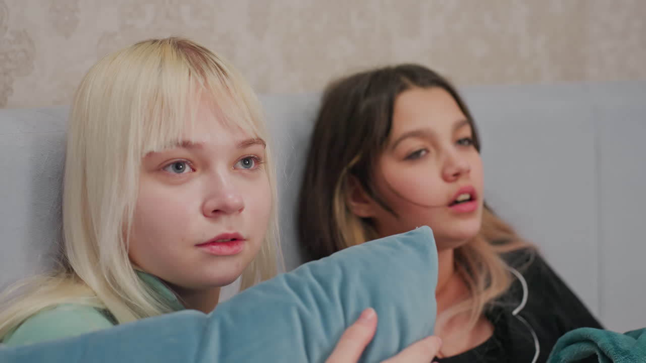 young ladies sitting close together on bed watching horror movie looking scared, one hiding behind pillow while other clutches blanket with tense expression