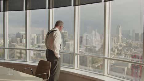an older businessman walks away from the flipover in his office and overlooks the skyline of the city of rotterdam, the netherlands from his high office window.