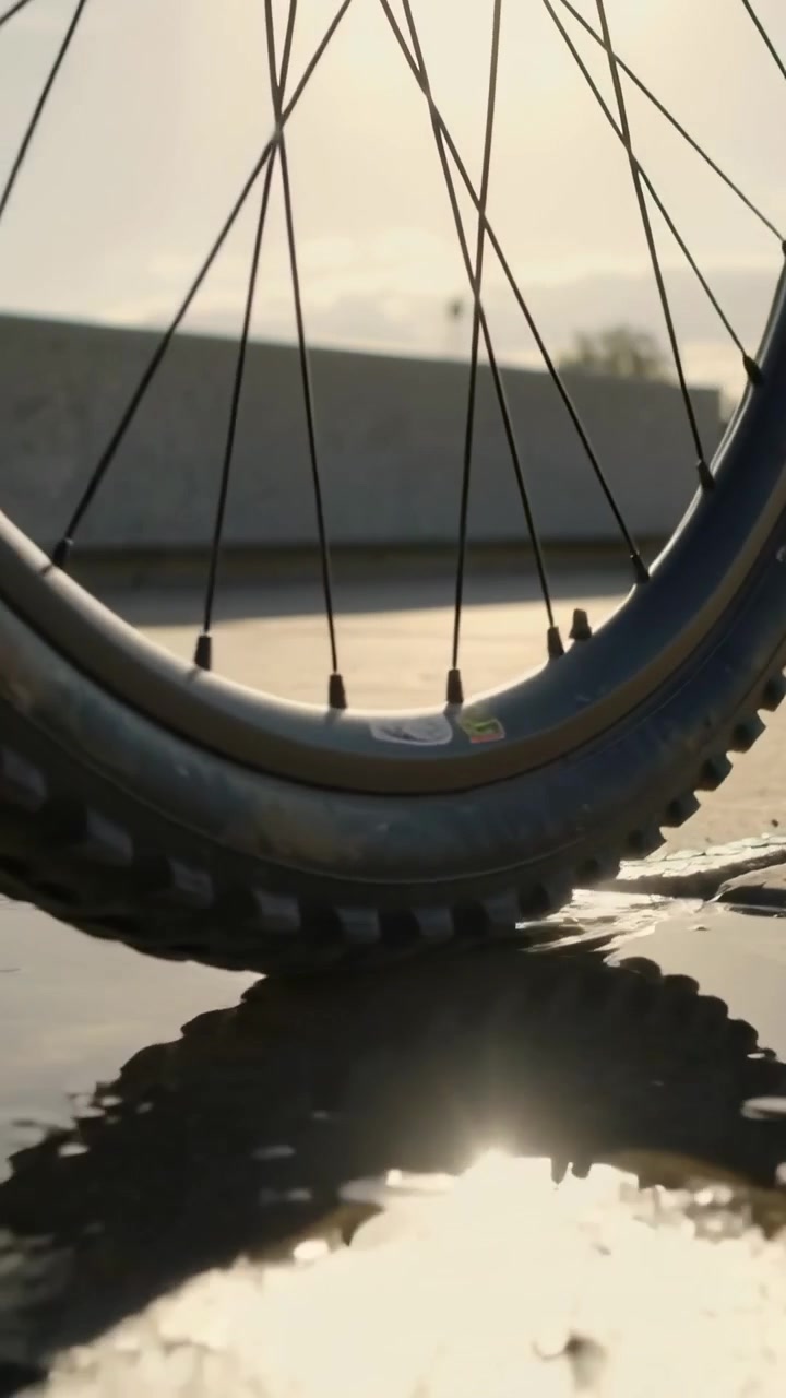 Video verticale: Ruota di bicicletta che rotola dopo una spinta, comprimendosi sul marciapiede soleggiato e schizzando una pozzanghera.