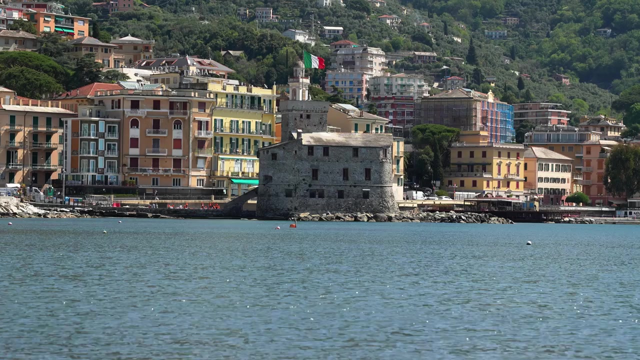Groothoekopname van het iconische kasteel van Rapallo tegen kleurrijke Ligurische gebouwen, met een helderblauwe zee op de voorgrond en weelderige heuvels daarachter, onder helder natuurlijk daglicht, geen mensen zichtbaar.