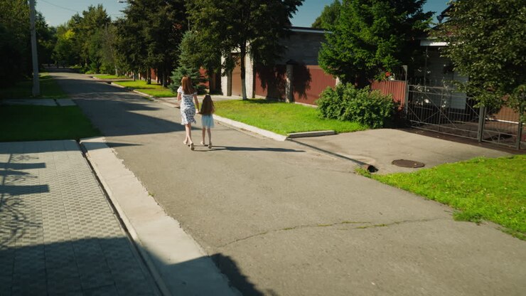 Rear view of woman in floral dress and young girl in light blue dress running hand in hand along quiet sunny residential street, surrounded by green grass, trees, fences, and paved sidewalks