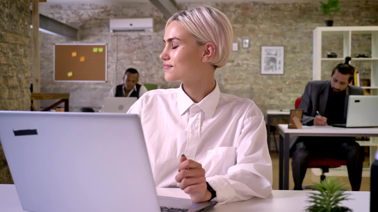 Giovane e felice donna d'affari seduta al laptop in ufficio, che balla, mentre i colleghi fanno networking con le tecnologie; concetto di lavoro, relax e comunicazione.