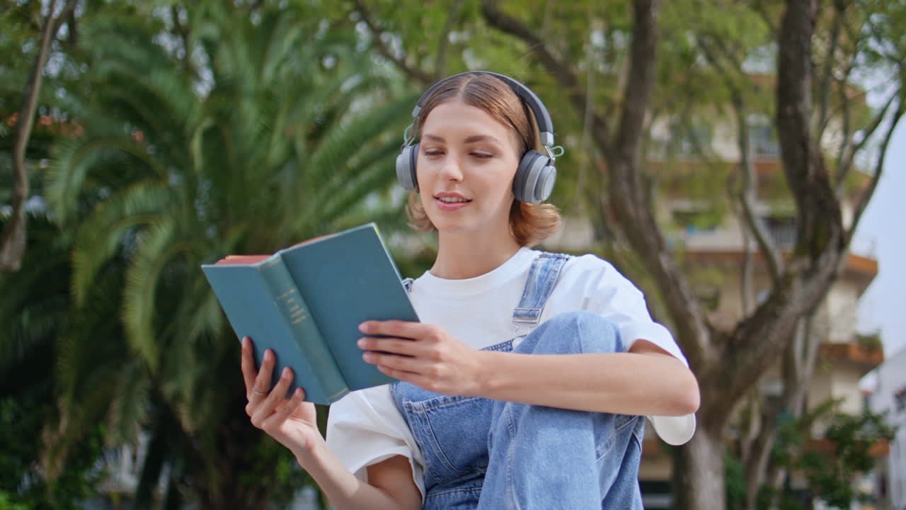 Calm girl reading novel listening music in headphones at sunny street closeup