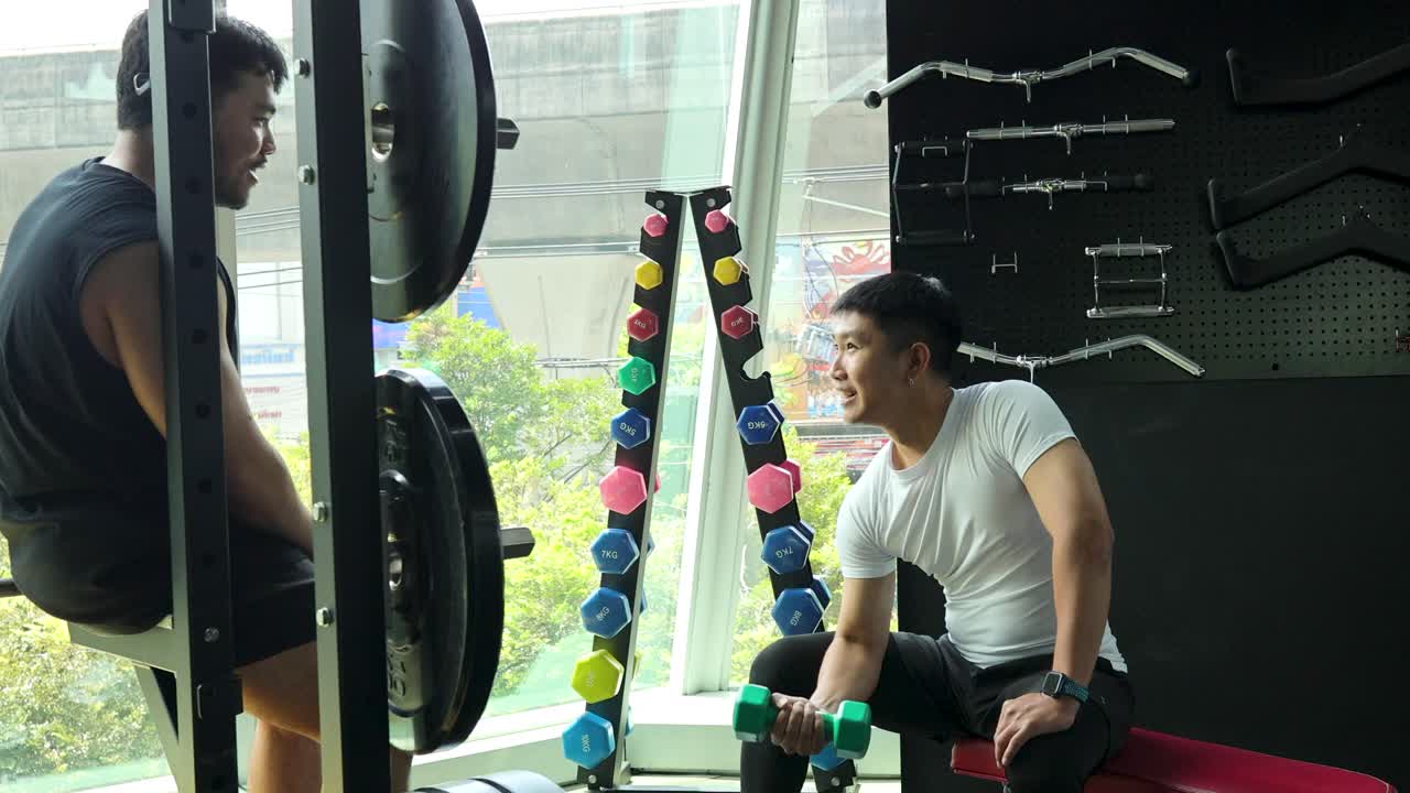 Two men converse beside gym equipment, one seated and lifting a dumbbell. Natural daylight fills the modern fitness center, with a relaxed, friendly mood